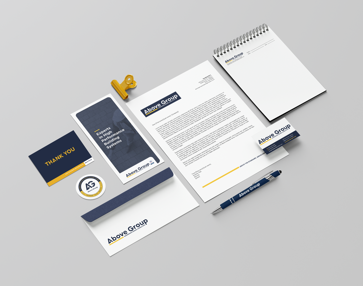 Mock-up of Above Group branded stationery items including a notepad, coaster, pen, envelope, and business card, arranged neatly on a grey background